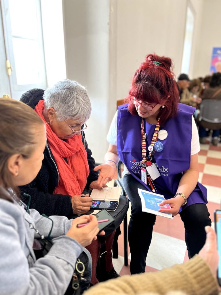 Mujer con pechera color morado, explicando a adulta mayor el funcionamiento de app en un teléfono